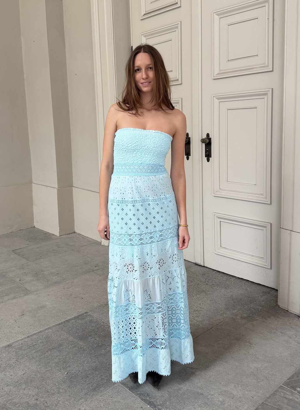 Woman standing outdoors in a light blue strapless maxi dress with lace panels, posing in front of white paneled doors.