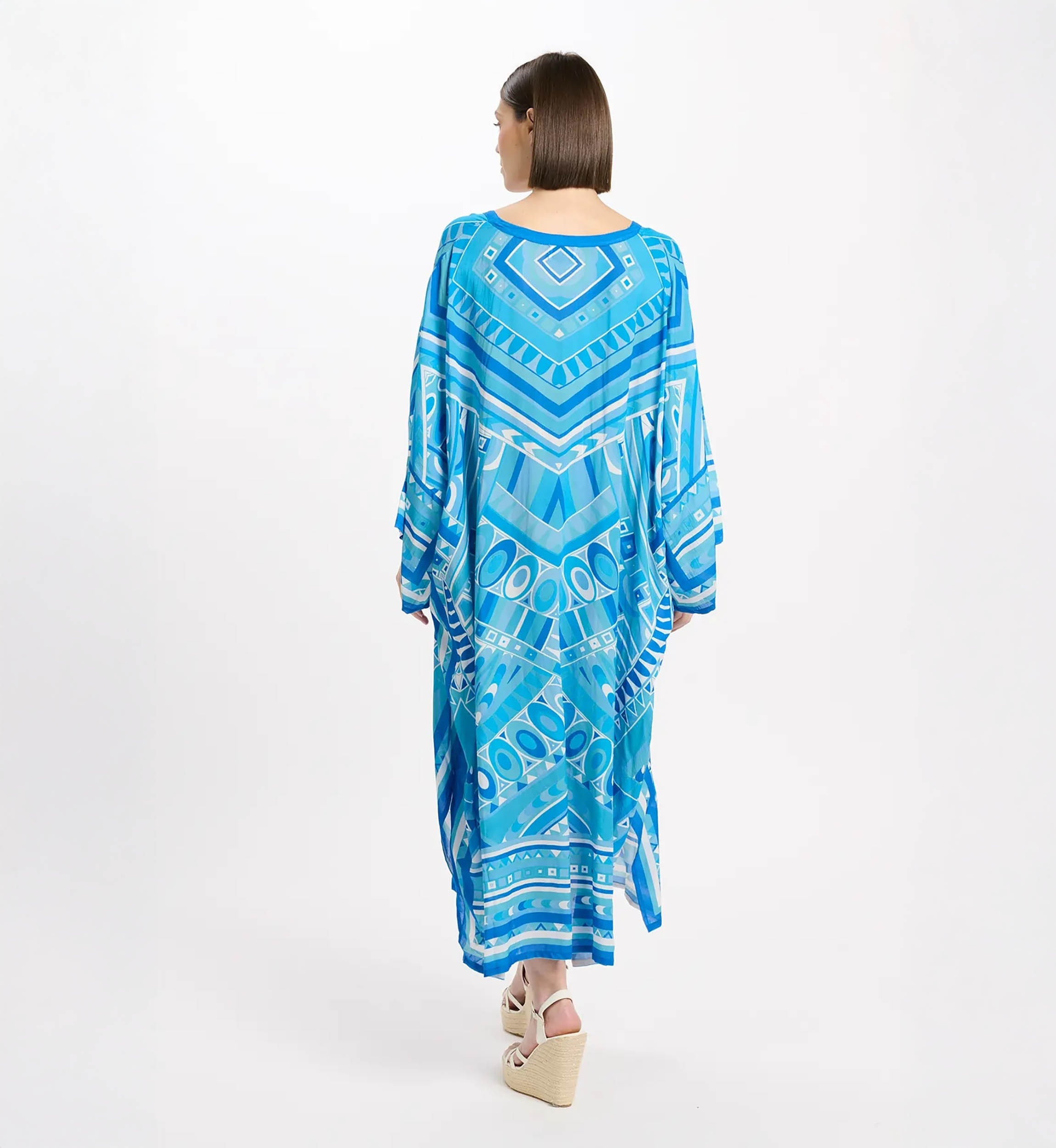 Back view of a woman in a long blue geometric-pattern maxi dress, standing against a white studio backdrop, wearing wedge sandals.