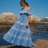 Woman in a light blue off-shoulder dress with white lace walking on rocky shore by the sea under a clear blue sky