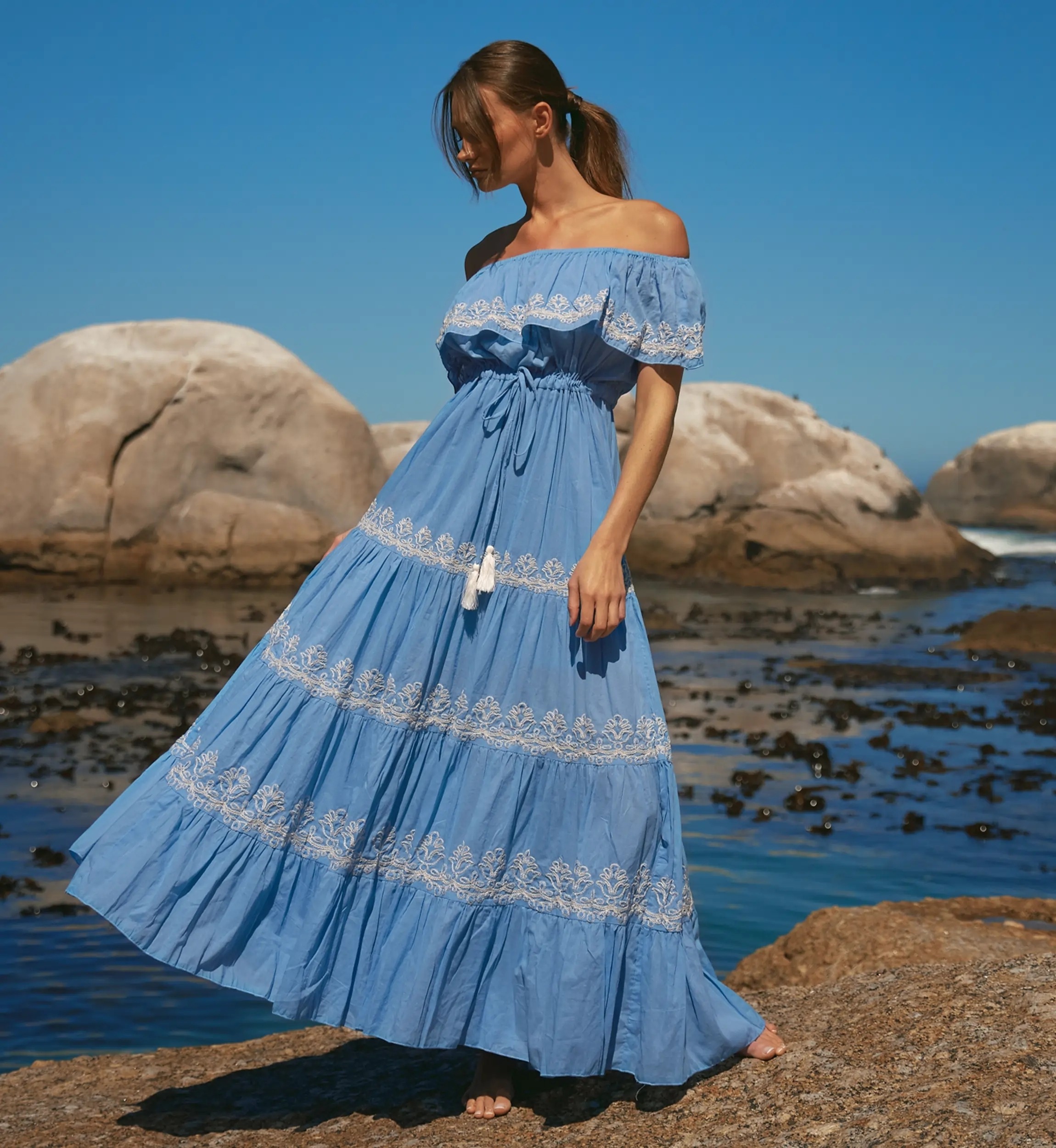 Woman in a light blue off-shoulder dress with white lace walking on rocky shore by the sea under a clear blue sky