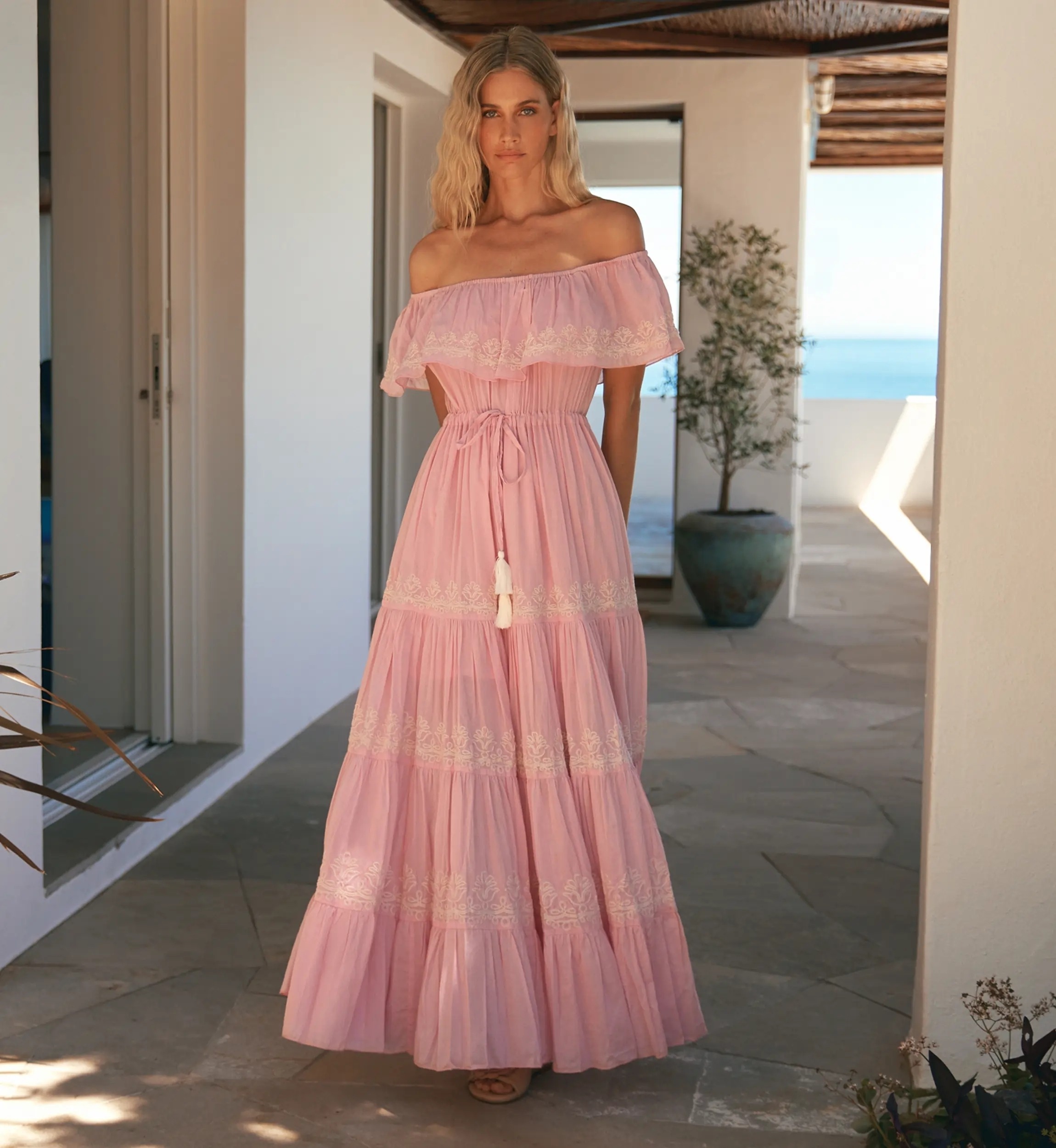 Woman in a pink off-shoulder maxi dress with lace trim, standing on a terrace with an ocean view.