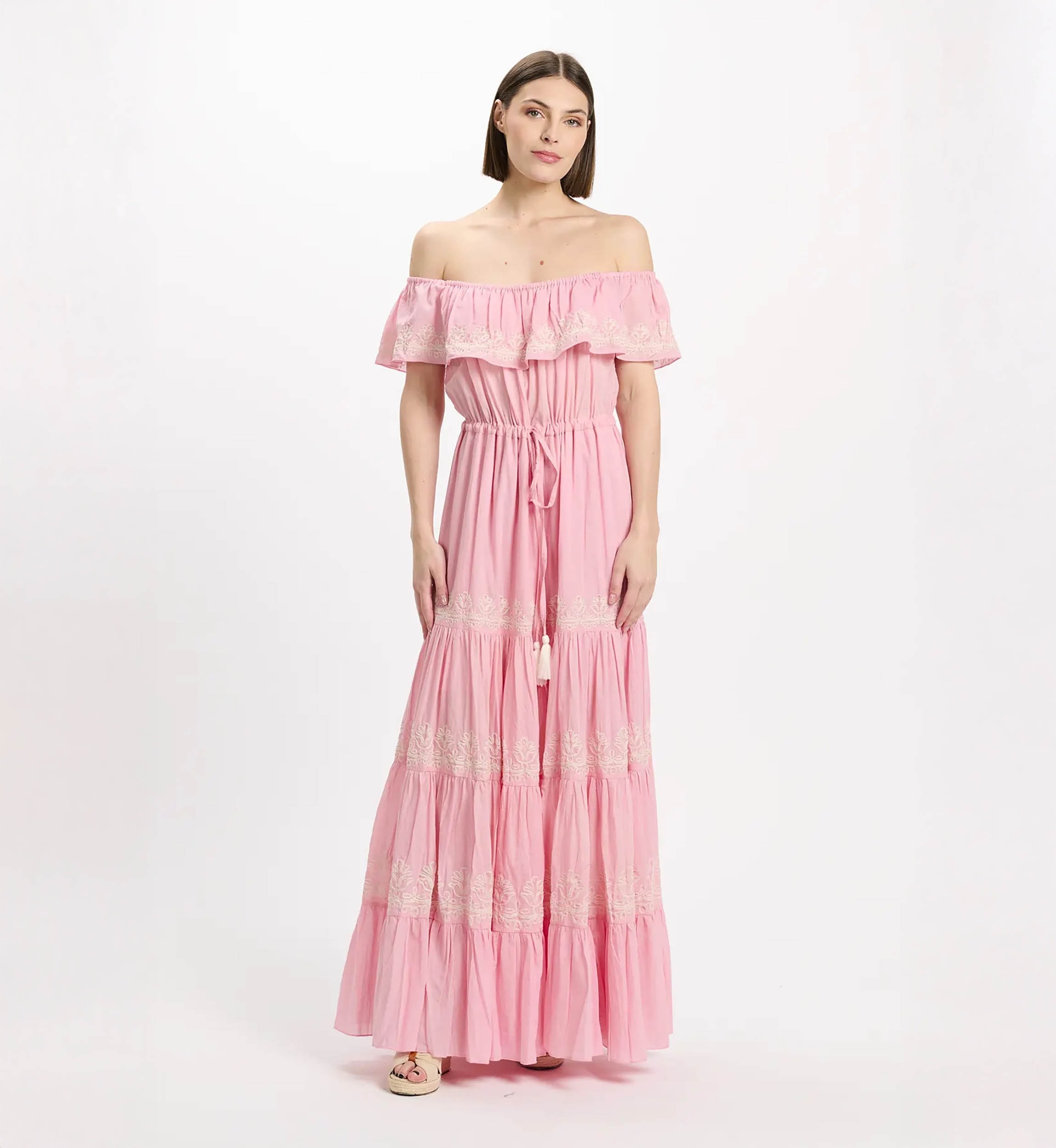 Pink off-the-shoulder maxi dress with a ruffled neckline and white lace trim, cinched waist, and tiered, lace-accented skirt on a model.