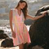 Woman in a pastel pink striped sundress leaning against a large rock on a sunny beach with waves in the background.