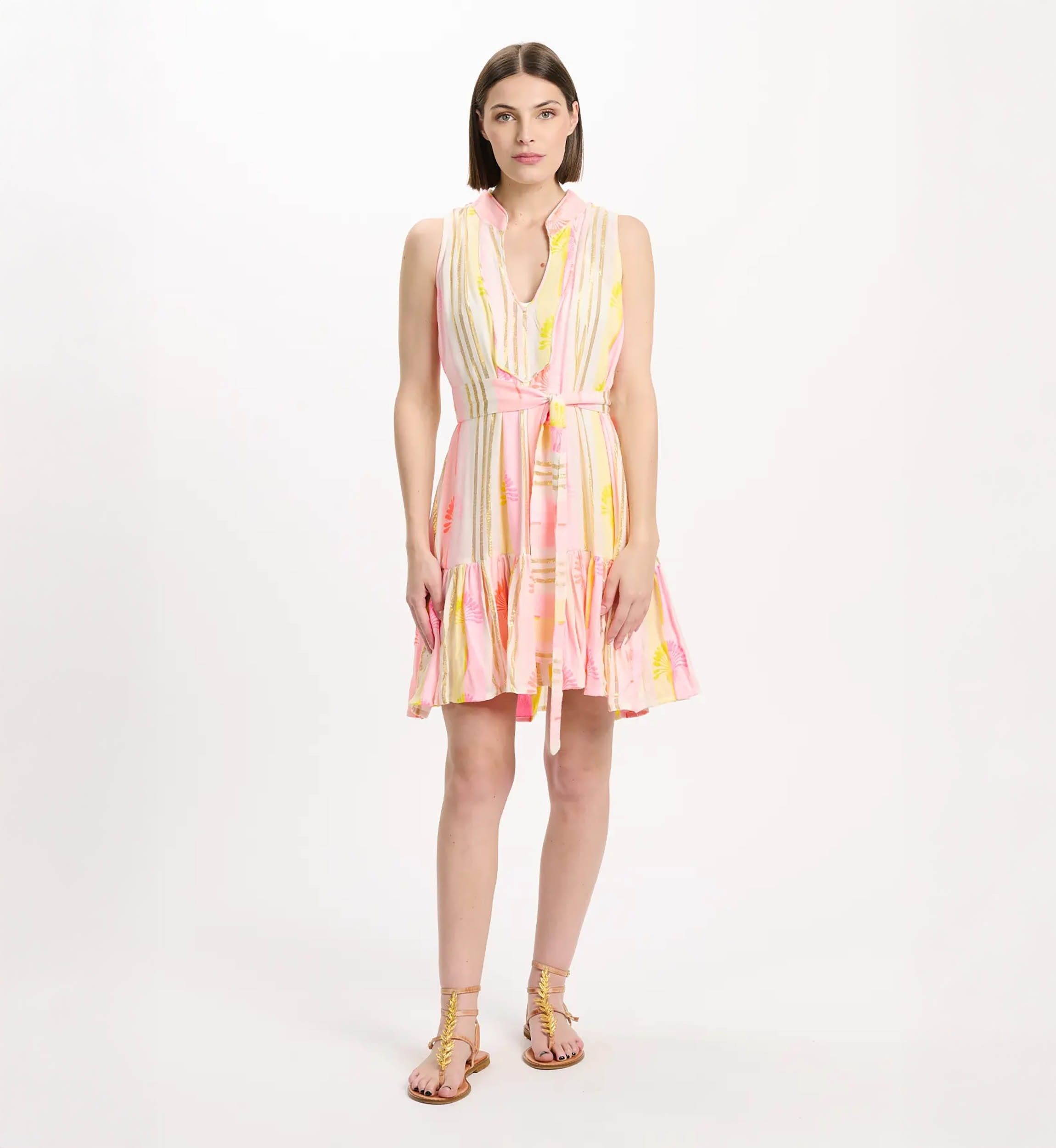 Model wearing a pink and yellow striped sleeveless sundress with a tied waist and sandals, standing against a white studio backdrop.