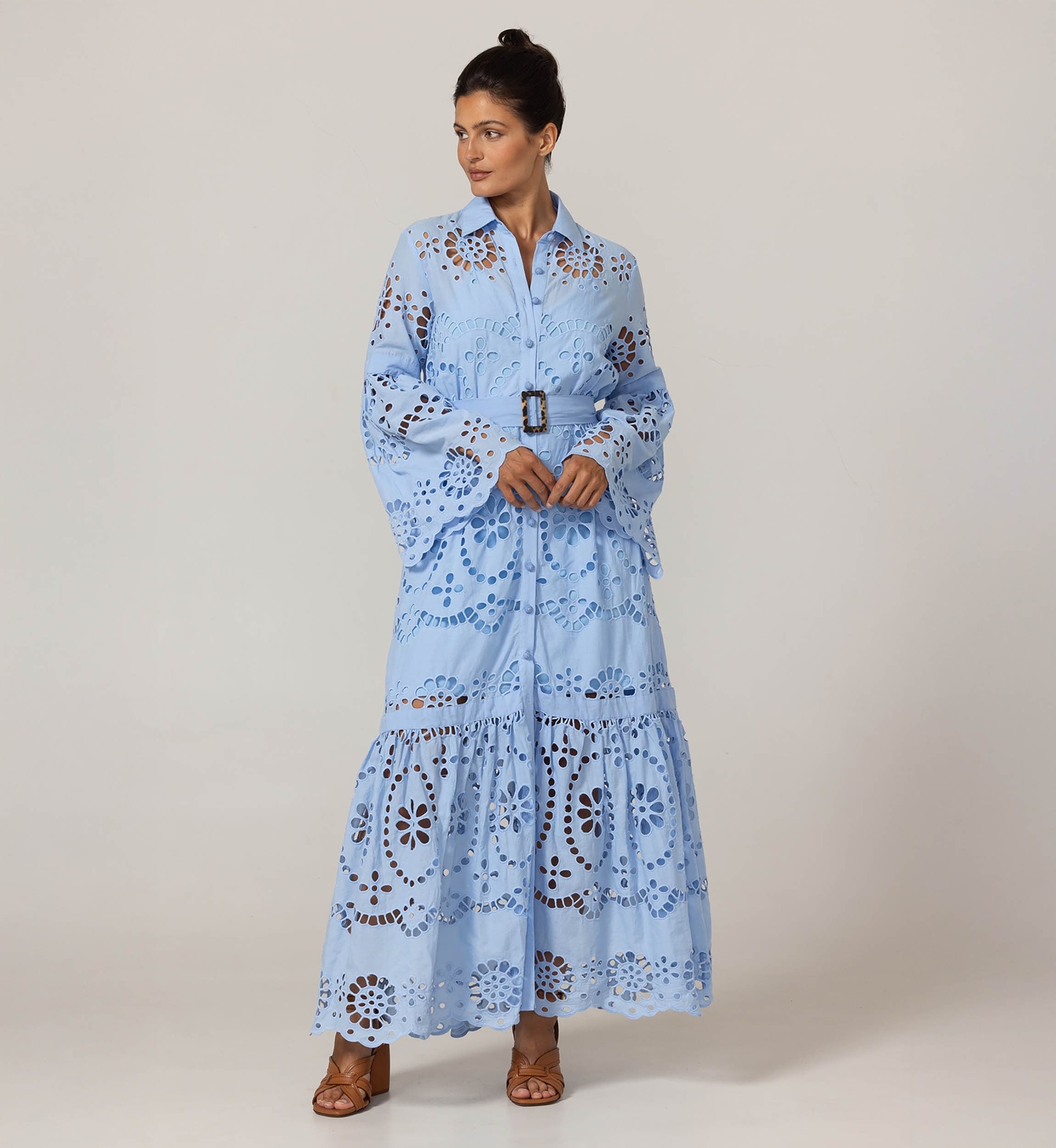 Woman in a light-blue, floor-length eyelet maxi dress with a belt, wide bell sleeves, and brown sandals against a neutral backdrop.