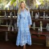 Woman in a light blue embroidered maxi dress with a belt, posing beside wooden shelves filled with carved statues outdoors.