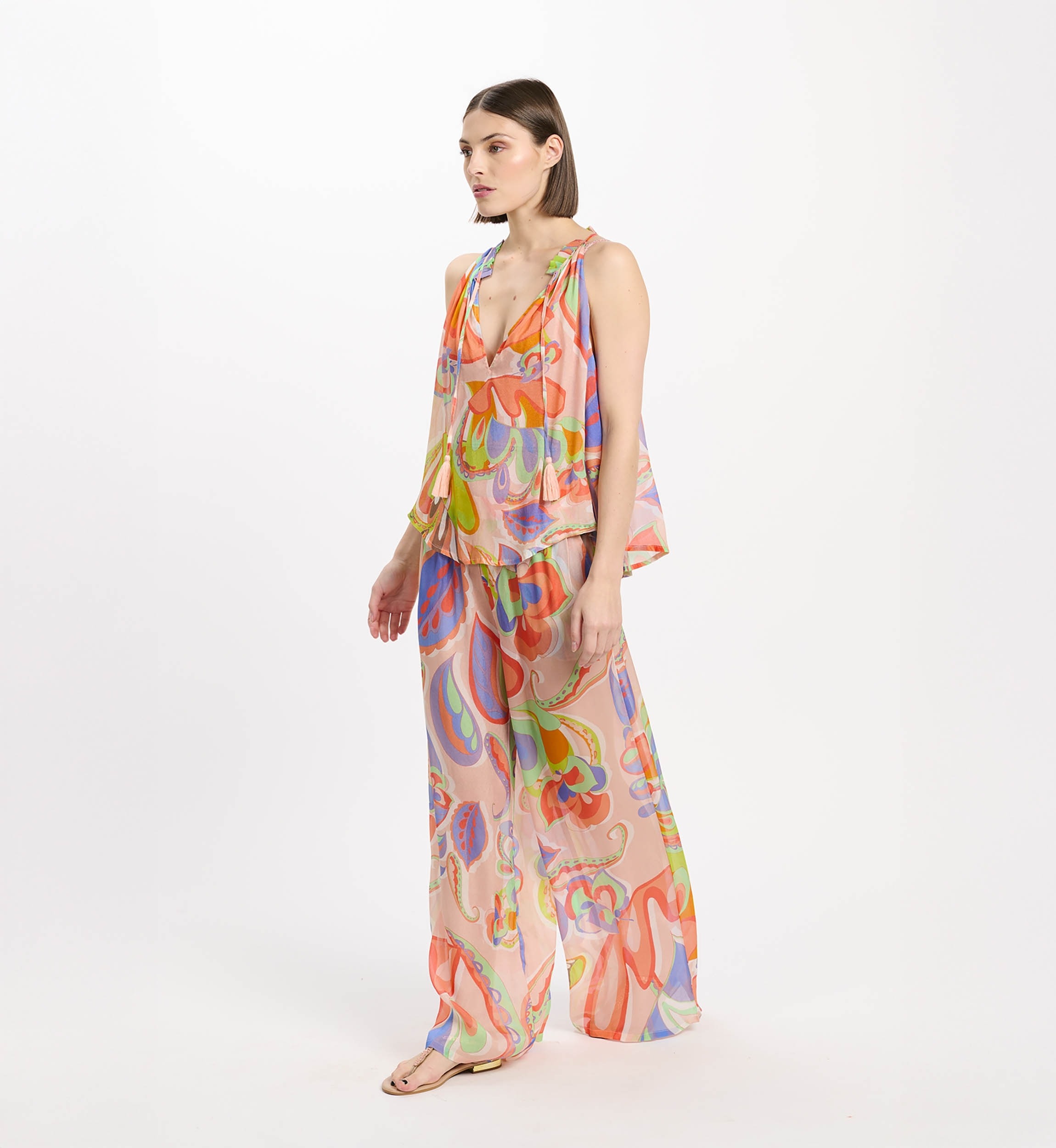 Model wearing a colorful, abstract-printed halter jumpsuit with wide legs on a white studio backdrop — peach and pastel tones dominate.