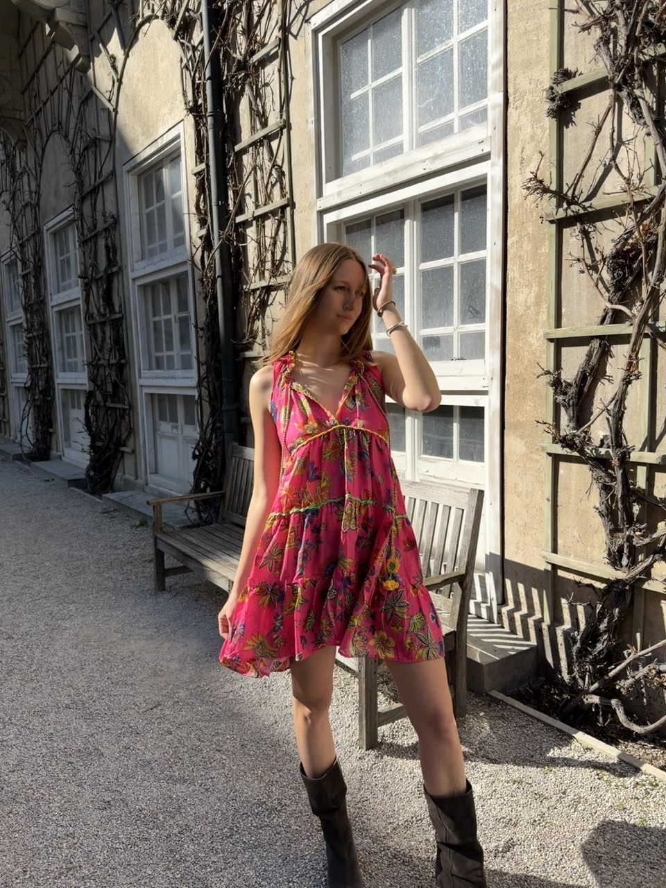 Woman in a bright pink floral sundress and brown knee-high boots posing on a gravel path beside an ivy-covered, sunlit building with a bench nearby.