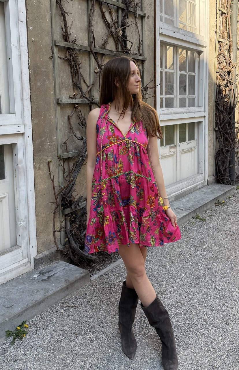 Woman in a bright pink floral sundress and black knee-high boots posing outdoors beside a vine-covered wall.