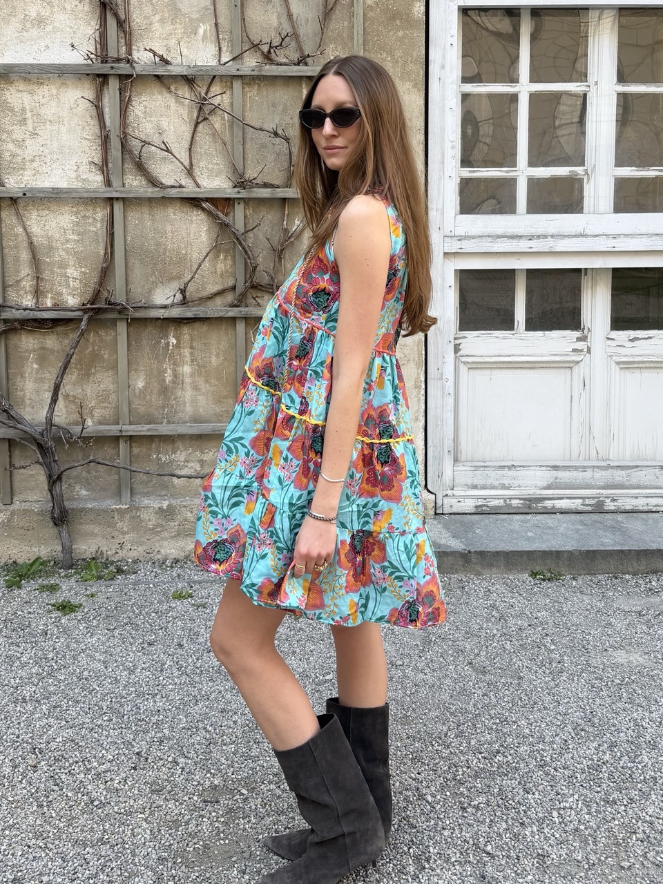Woman wearing a bright blue floral sundress, sunglasses, and knee-high dark boots, standing sideways outdoors beside a weathered wall with a window.