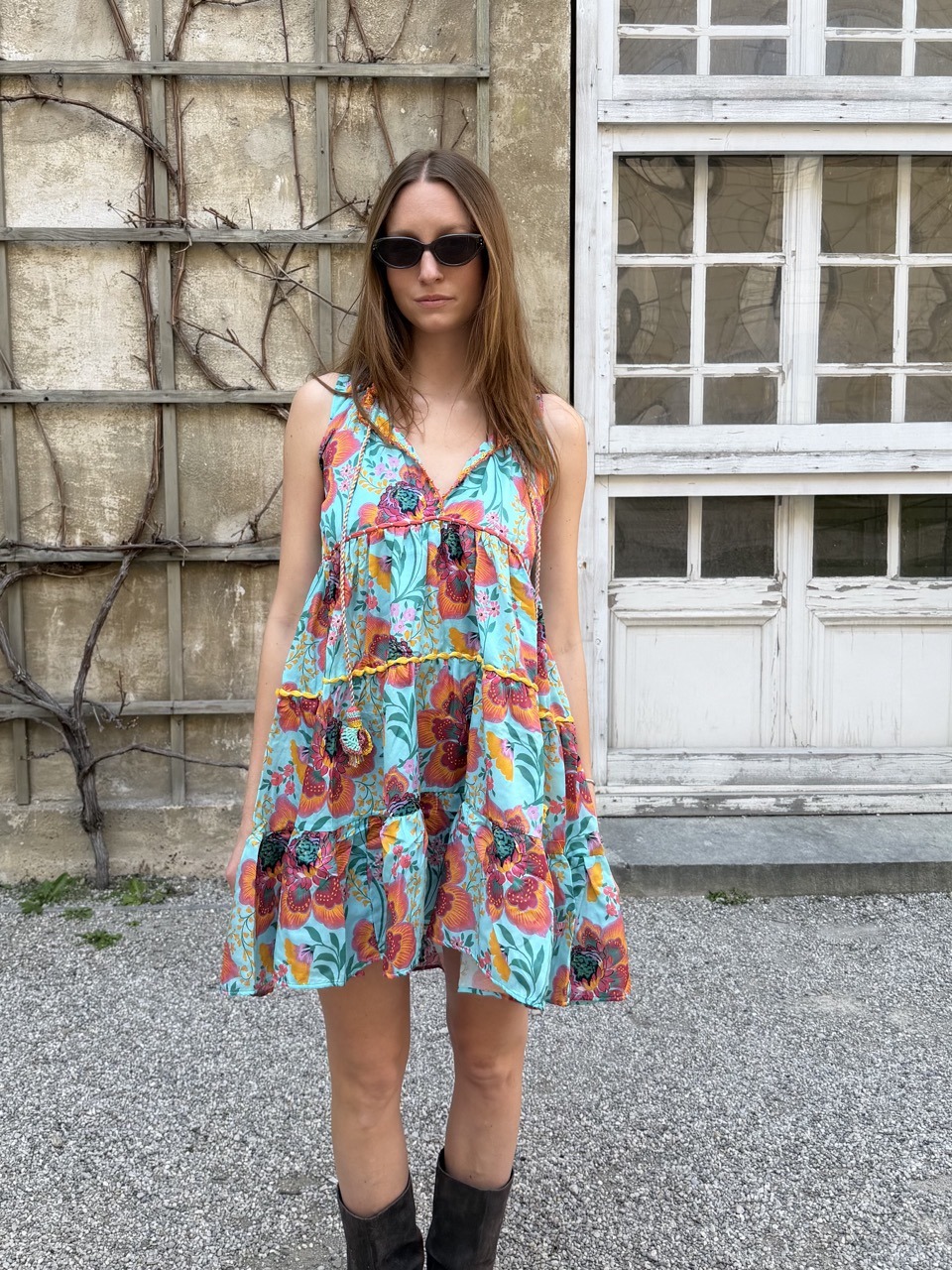 Woman wearing a bright turquoise floral dress and sunglasses stands outdoors beside an old wall and a windowed door.