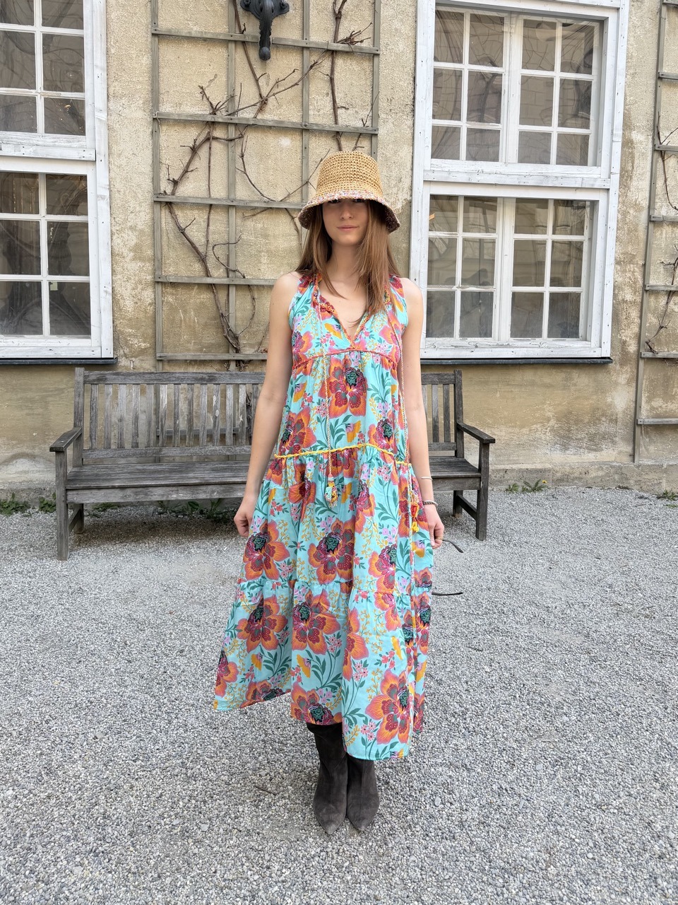 Woman in a turquoise floral sleeveless dress and straw hat stands on gravel in front of an old stone building with wooden benches.