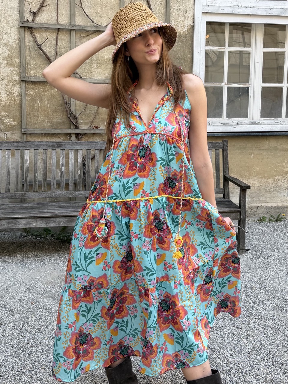 Woman in a turquoise floral sundress and a straw hat posing outdoors beside a wooden bench.