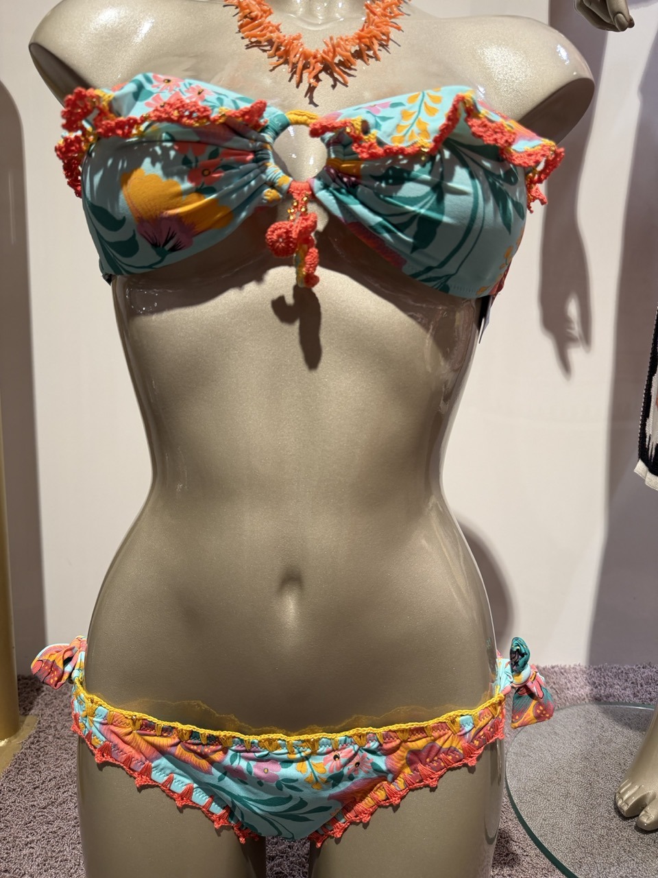 Turquoise tropical-print bikini with orange fringe on a display mannequin, with a coral necklace above the bust.