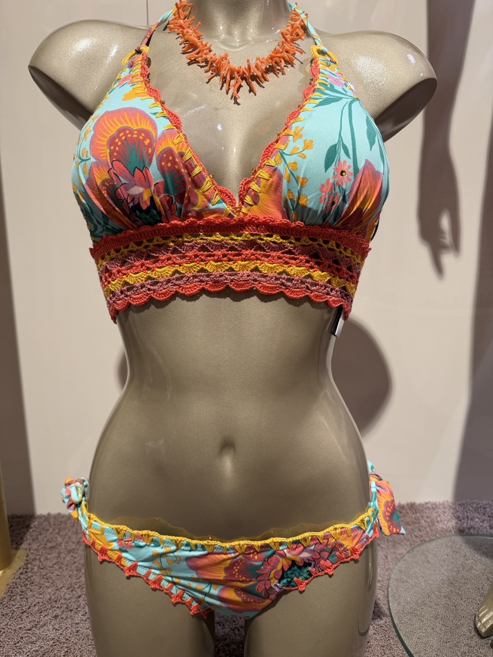 Tropical print bikini on a mannequin: turquoise top with orange-pink flowers and bright crochet trim, matching bottom with side ties and scalloped edges.