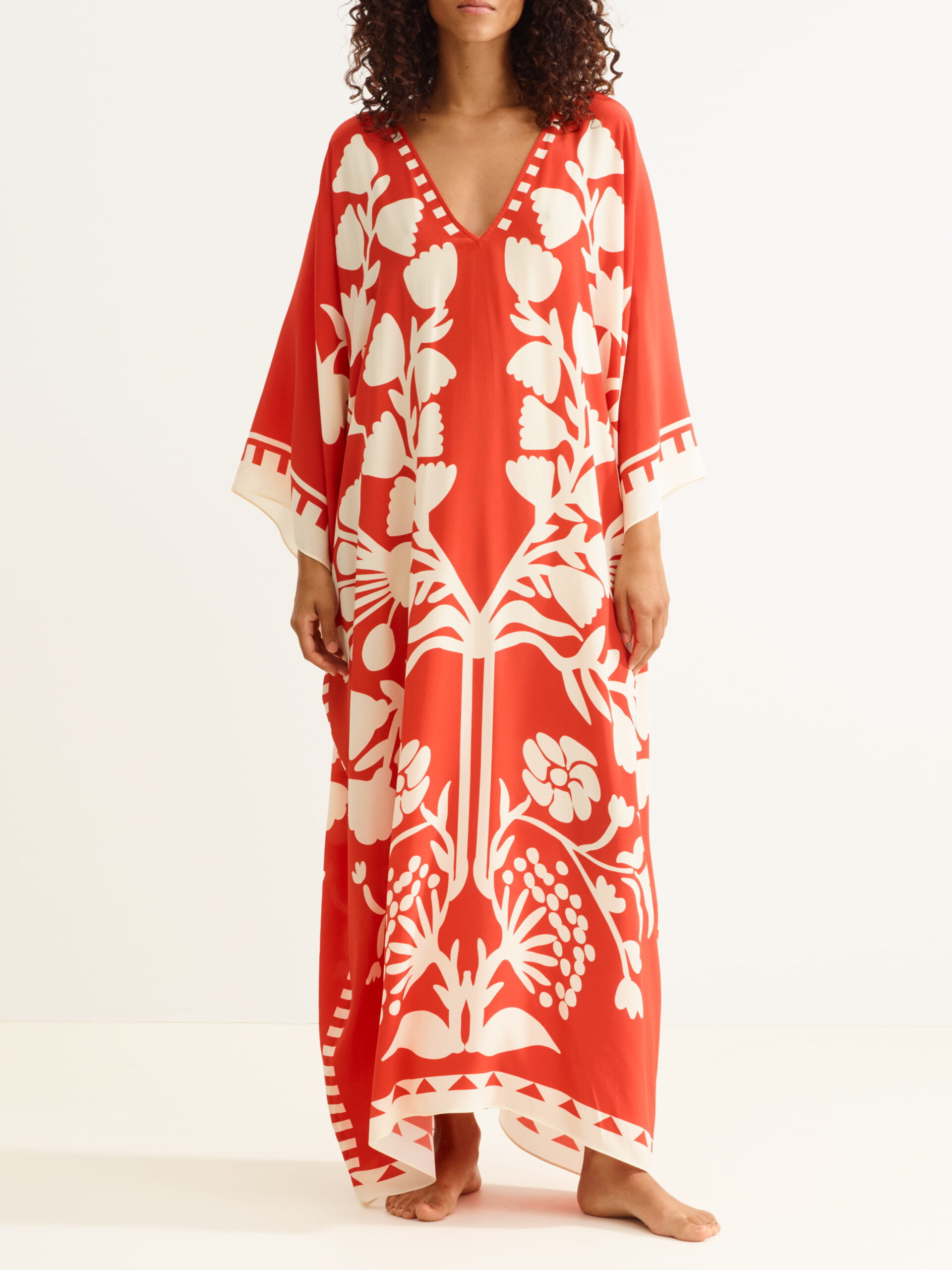 Woman wearing a flowing red kaftan with a bold white botanical print and a deep V-neck, standing barefoot.