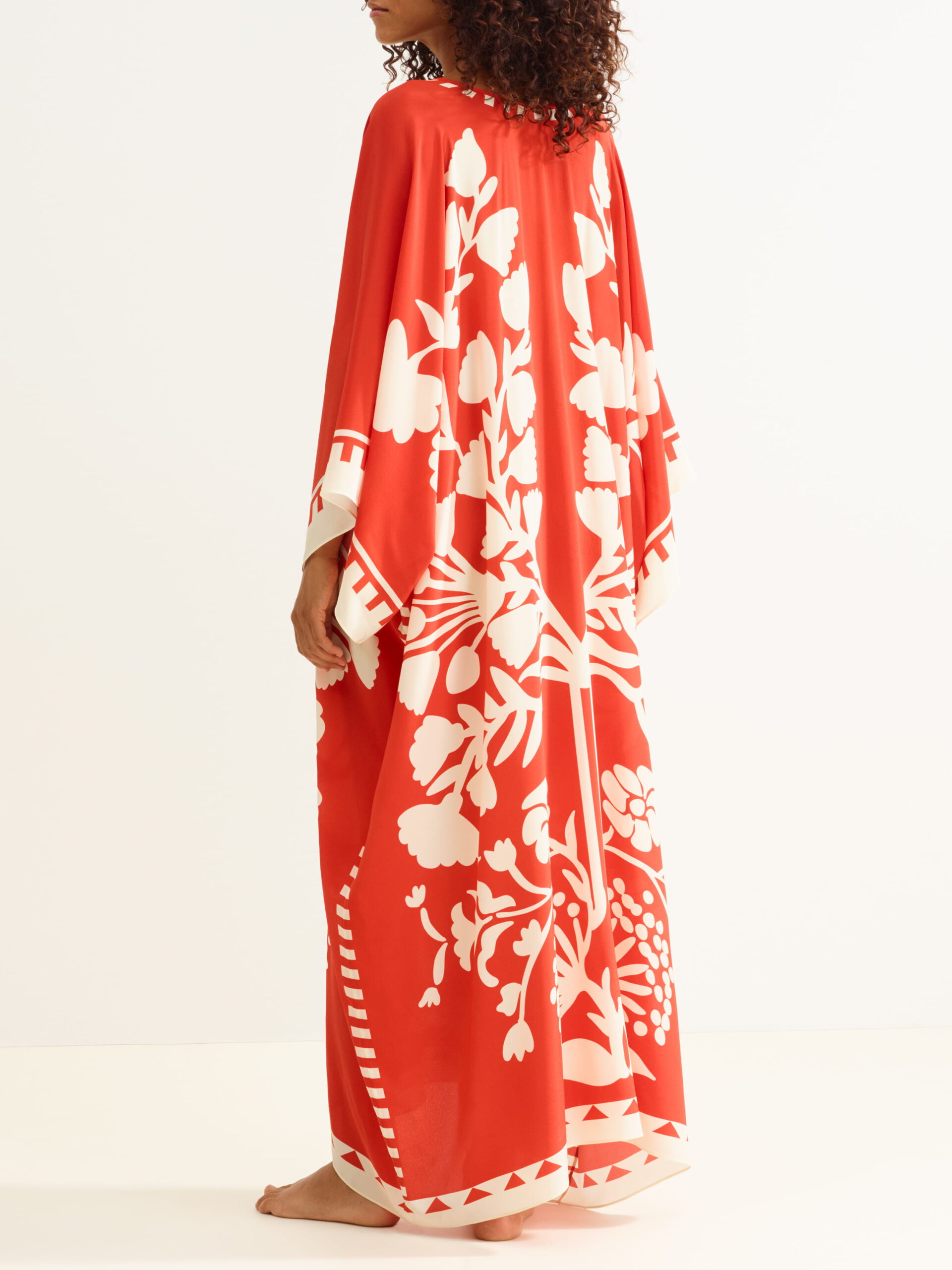 Back view of a person in a long red kaftan with bold white floral print, barefoot against a white background.