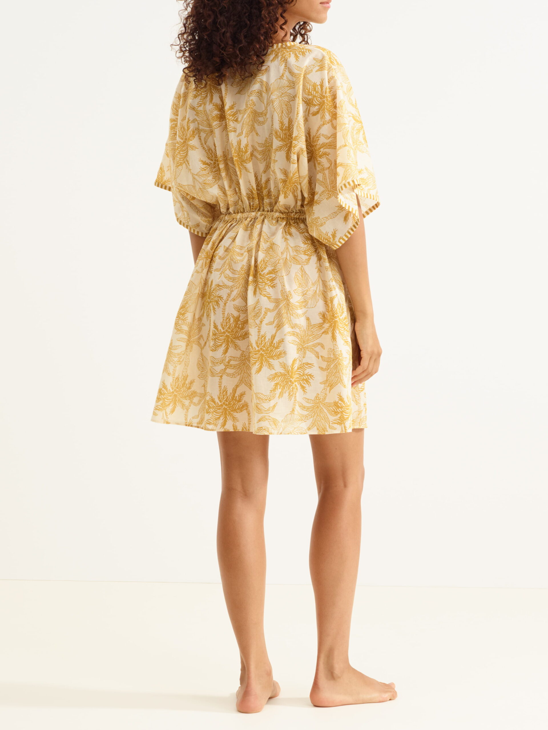 Back view of a person wearing a beige knee-length dress with a yellow palm-leaf print, elastic waist, and short flutter sleeves; barefoot on a light background.