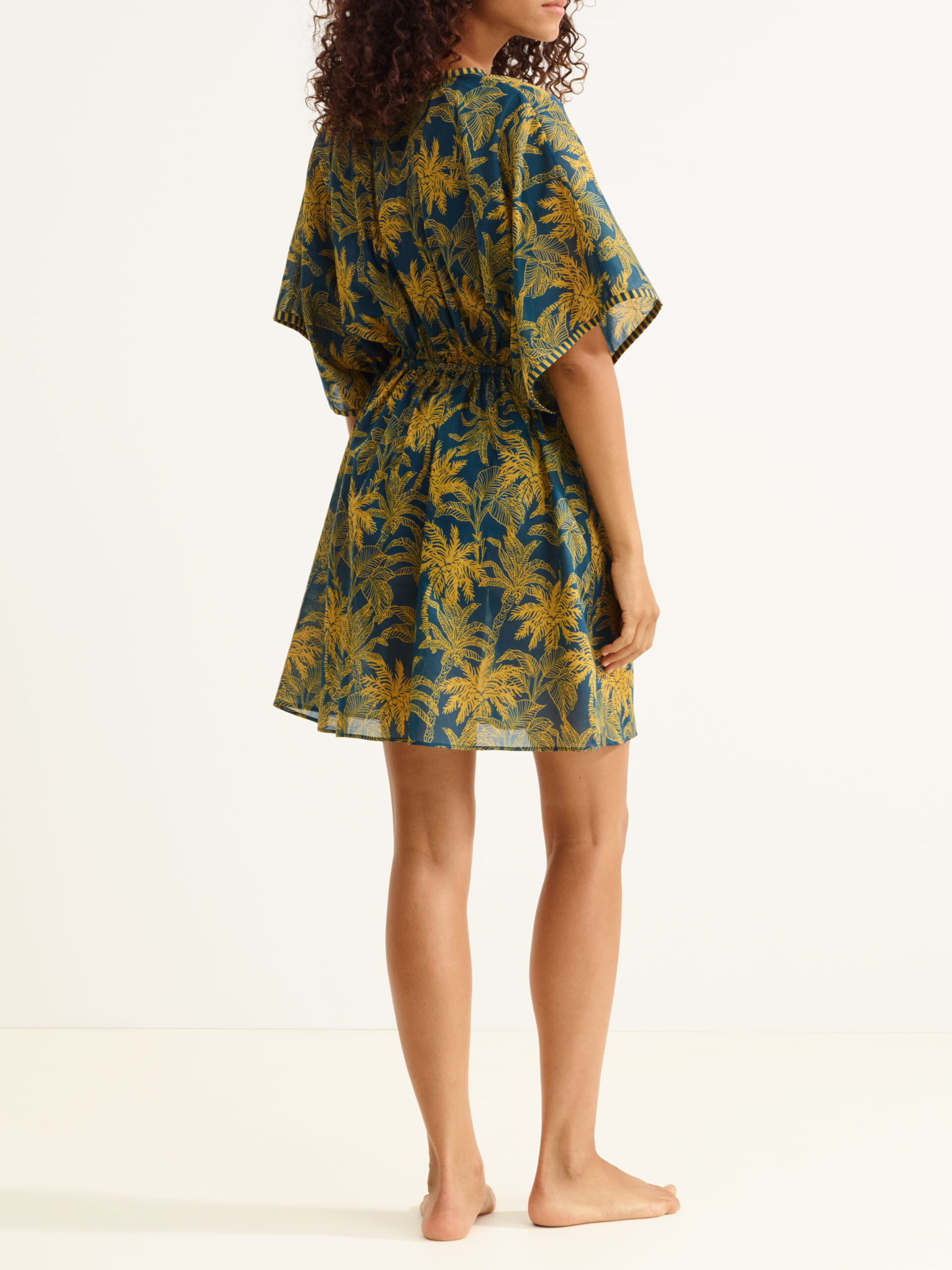 Woman standing in a teal-blue knee-length dress with a yellow palm-leaf pattern, viewed from the back.