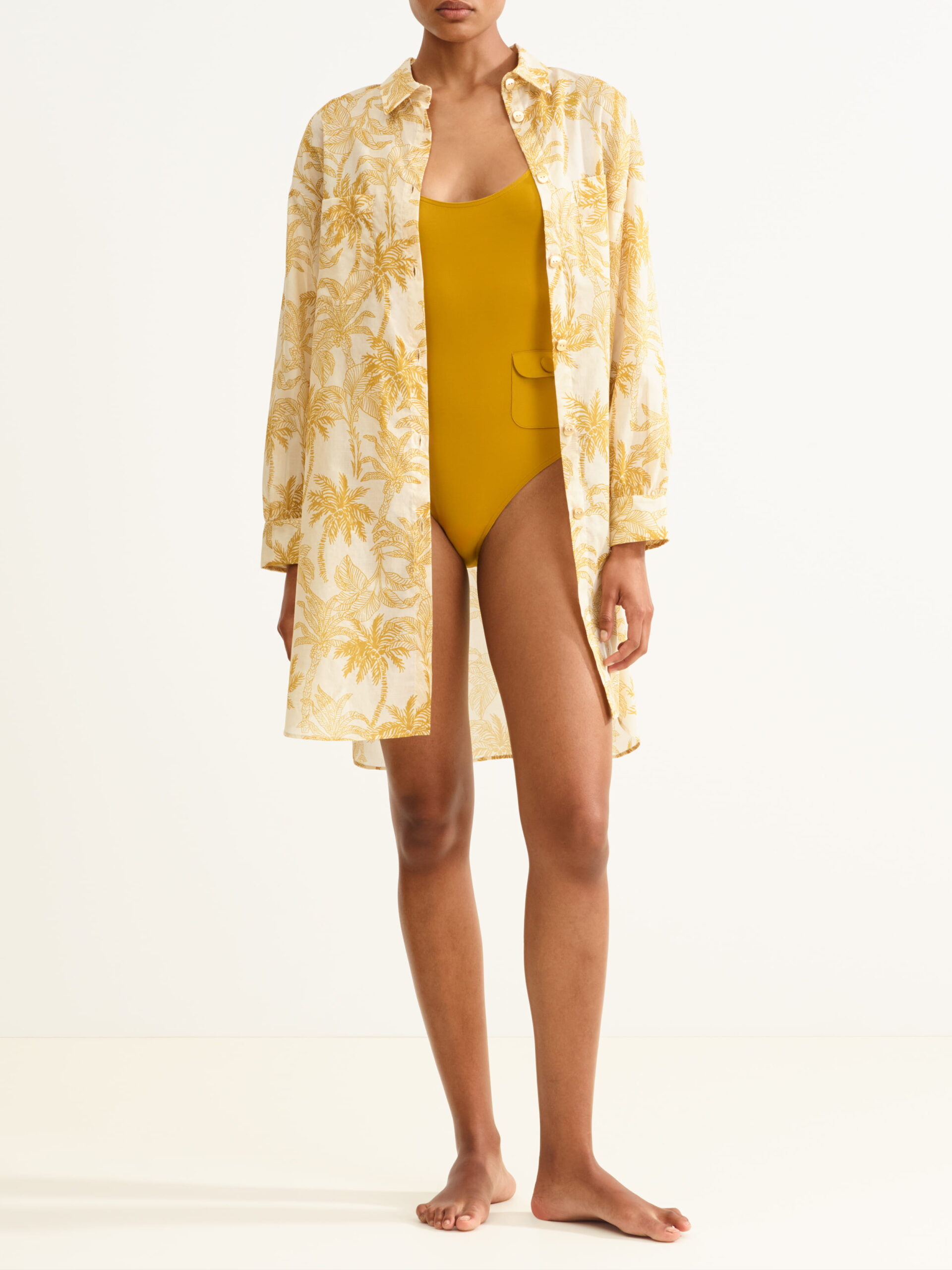 Model wearing a mustard-yellow one-piece swimsuit under a cream cover-up with a yellow palm print.