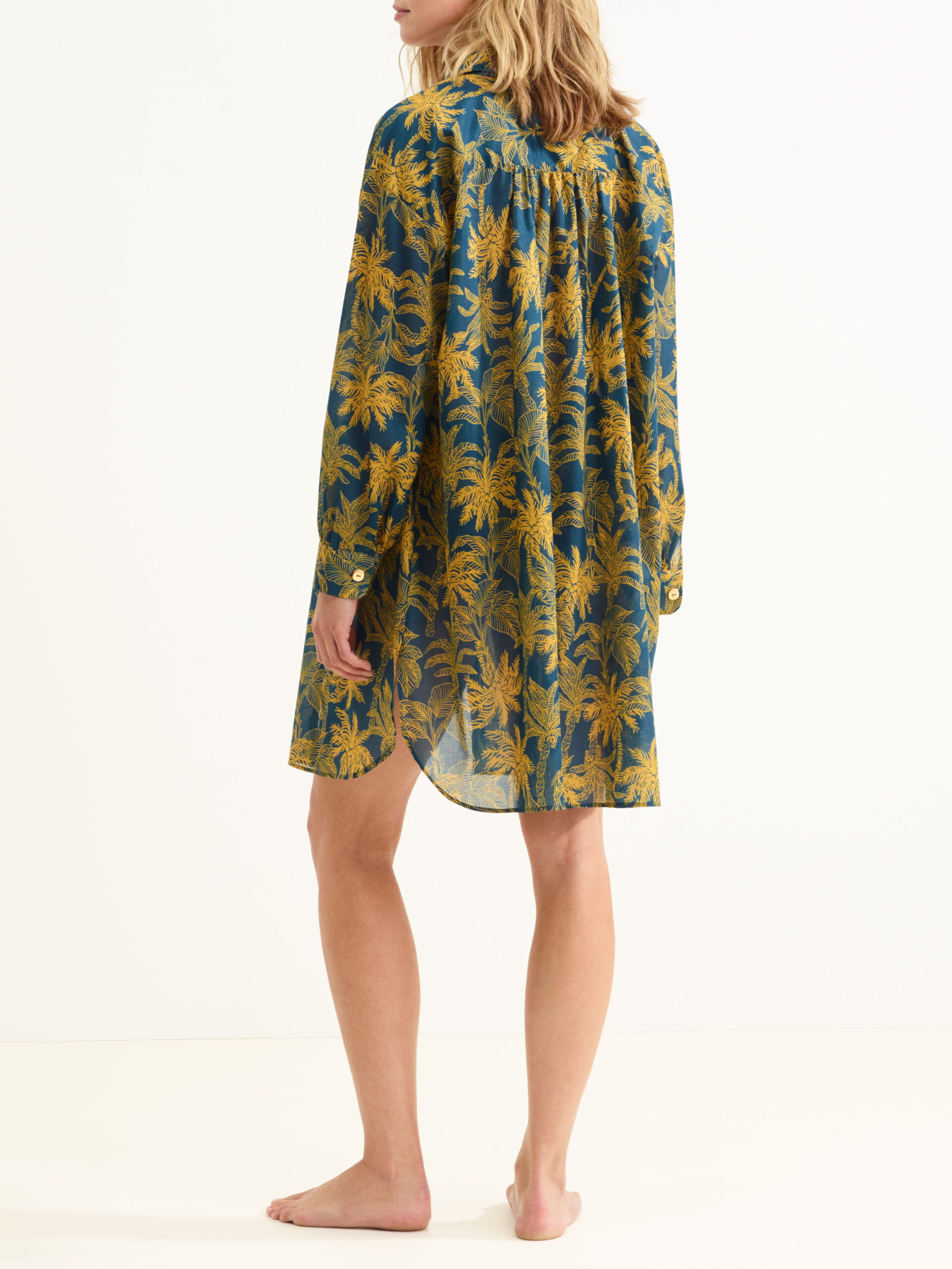 Back view of a woman wearing a loose blue dress with a yellow palm leaf print, knee-length and sheer.
