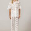 Model wearing a white eyelet lace shirt and matching wide-leg pants with circular floral cutouts, standing against a neutral backdrop, wearing light sandals.