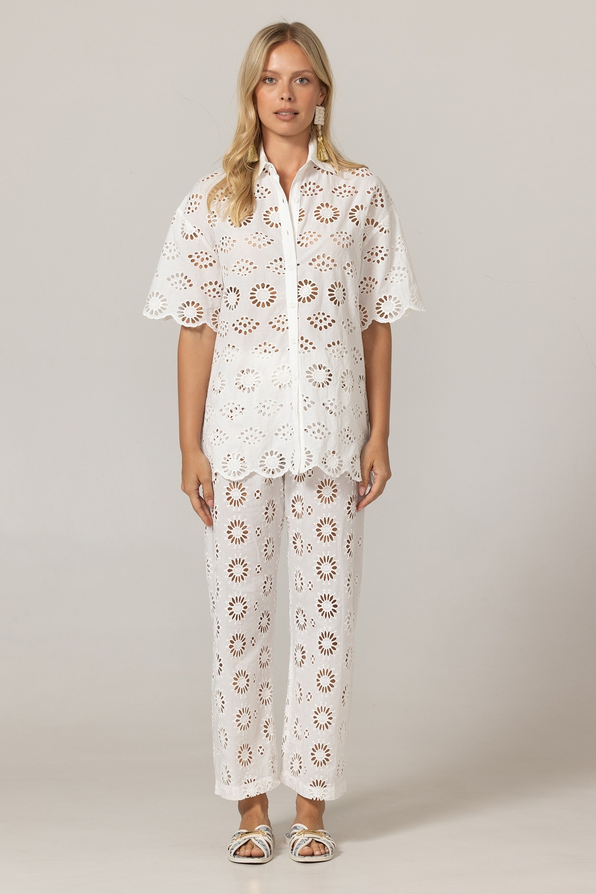 Model wearing a white eyelet lace shirt and matching wide-leg pants with circular floral cutouts, standing against a neutral backdrop, wearing light sandals.