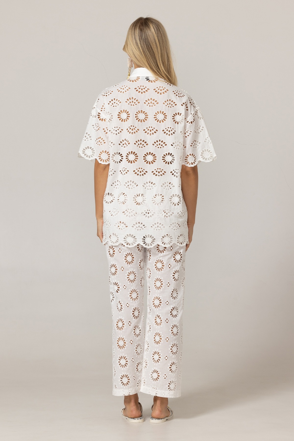 Back view of a woman wearing a white eyelet lace top and matching wide-leg pants against a neutral backdrop.