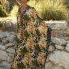 Woman wearing a colorful tropical palm-pattern kaftan, standing barefoot beside a stone wall outdoors in sunlight.