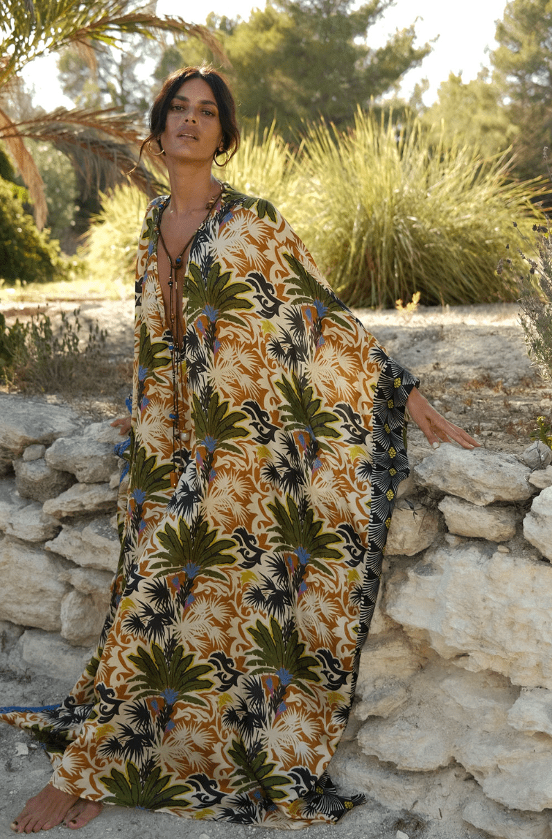 Woman wearing a colorful tropical palm-pattern kaftan, standing barefoot beside a stone wall outdoors in sunlight.