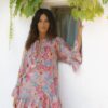 Woman in a pink floral dress leaning against a white wall beside a small window with green ivy above.