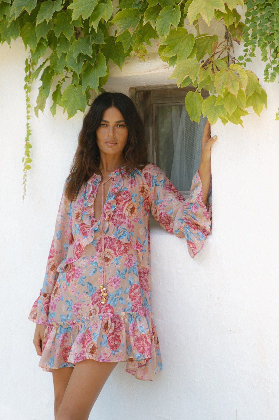 Woman in a pink floral dress leaning against a white wall beside a small window with green ivy above.