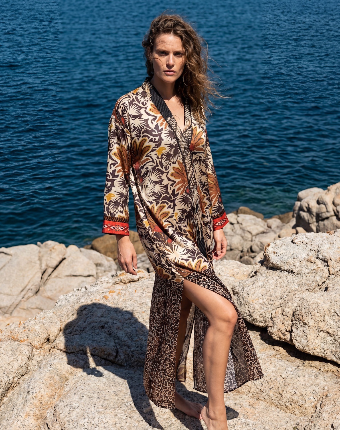 Woman standing barefoot on rocky shoreline by deep blue water, wearing a long tropical-patterned kimono with leopard-print hem and red cuffs.