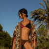 Woman in a red-and-black bikini with a patterned cover-up standing outdoors in bright sunlight, palm trees in the background.