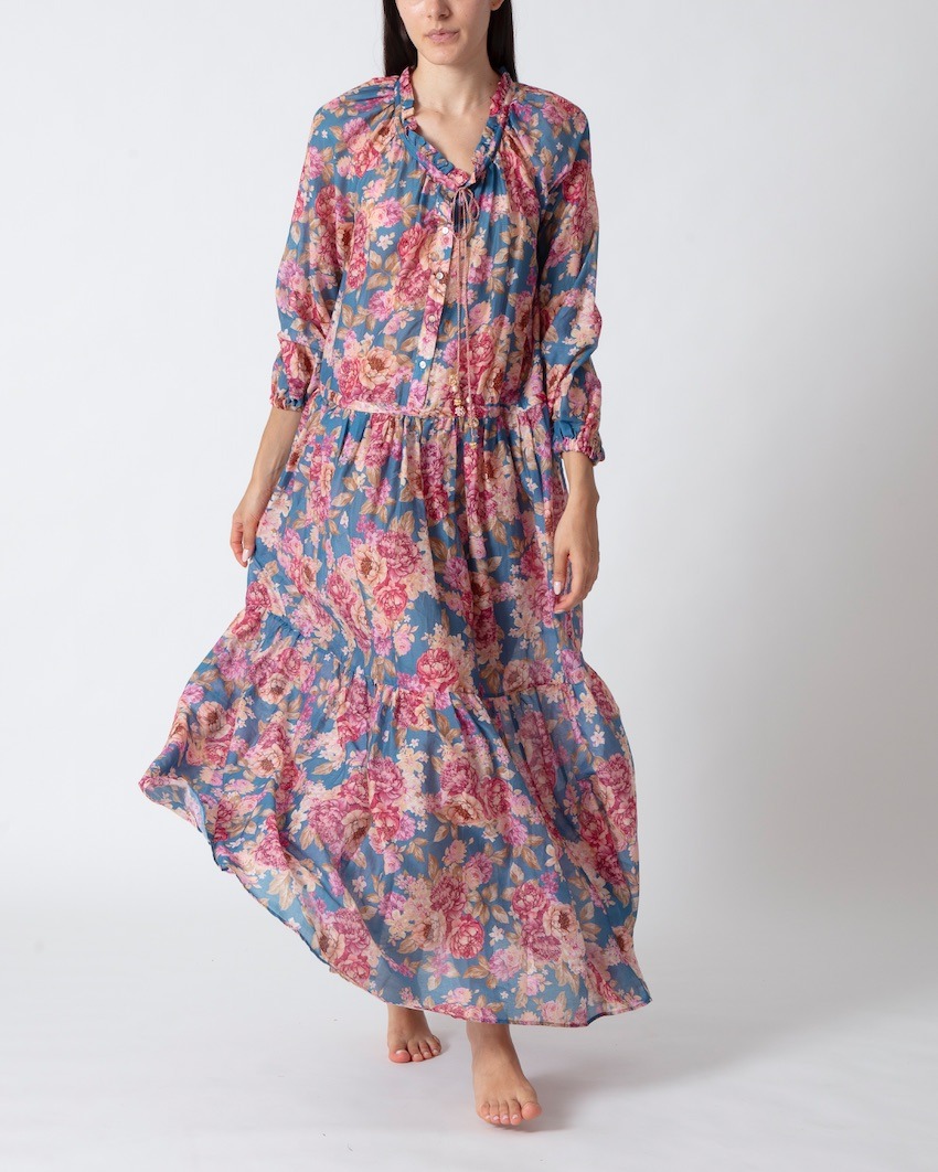 Woman wearing a blue floral maxi dress with pink blossoms, gathered waist, and flowing skirt, standing barefoot on white background.