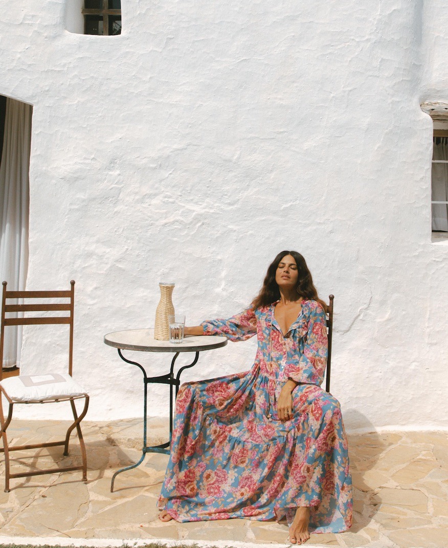 Woman in a blue and pink floral maxi dress sits barefoot at a round table on a sunny patio against a white stucco wall, eyes closed in relaxation.
