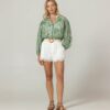 Full-body shot of a woman wearing a green botanical-print blouse, white eyelet shorts with a tan belt, and brown sandals, standing against a neutral backdrop.