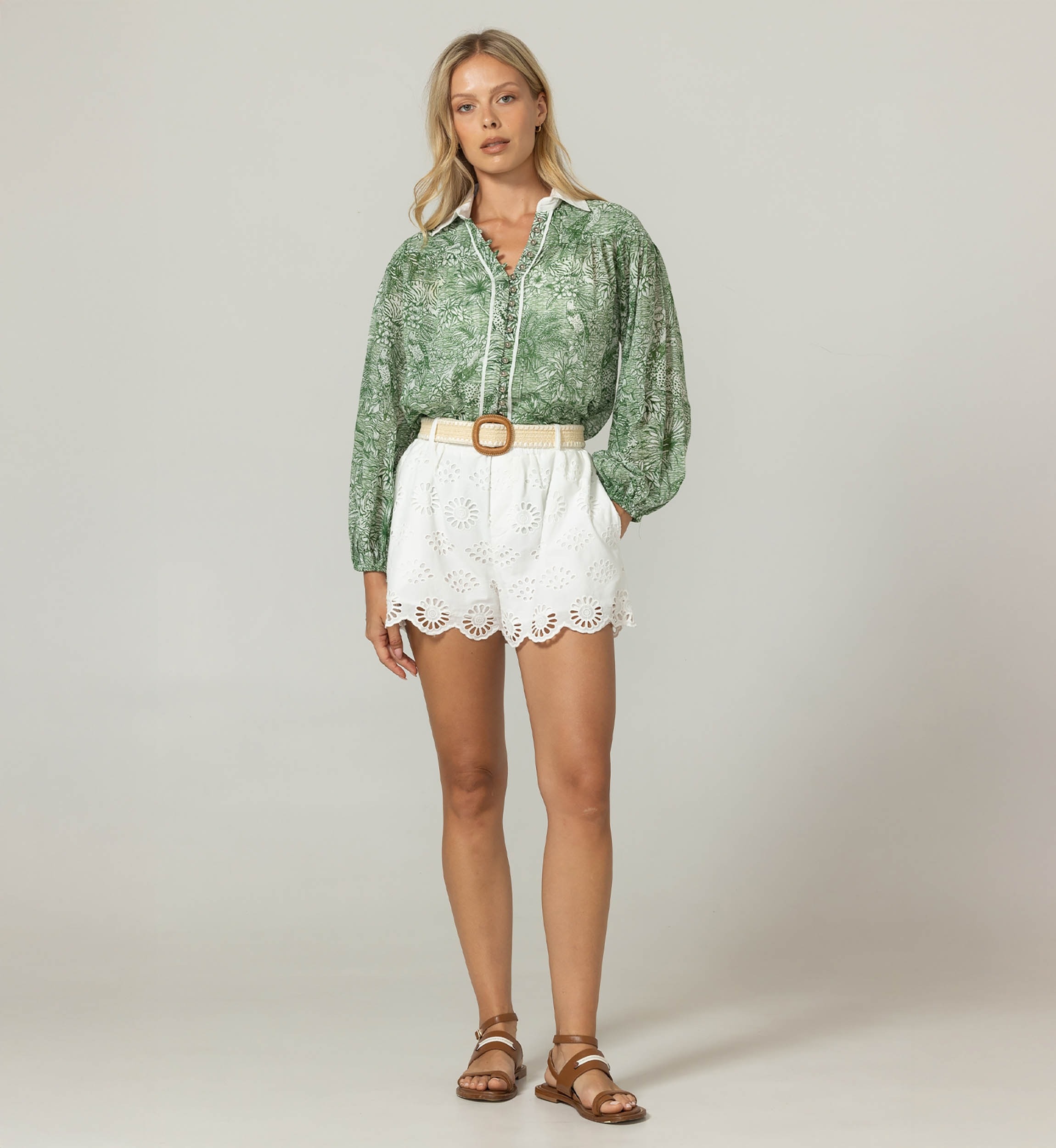 Full-body shot of a woman wearing a green botanical-print blouse, white eyelet shorts with a tan belt, and brown sandals, standing against a neutral backdrop.