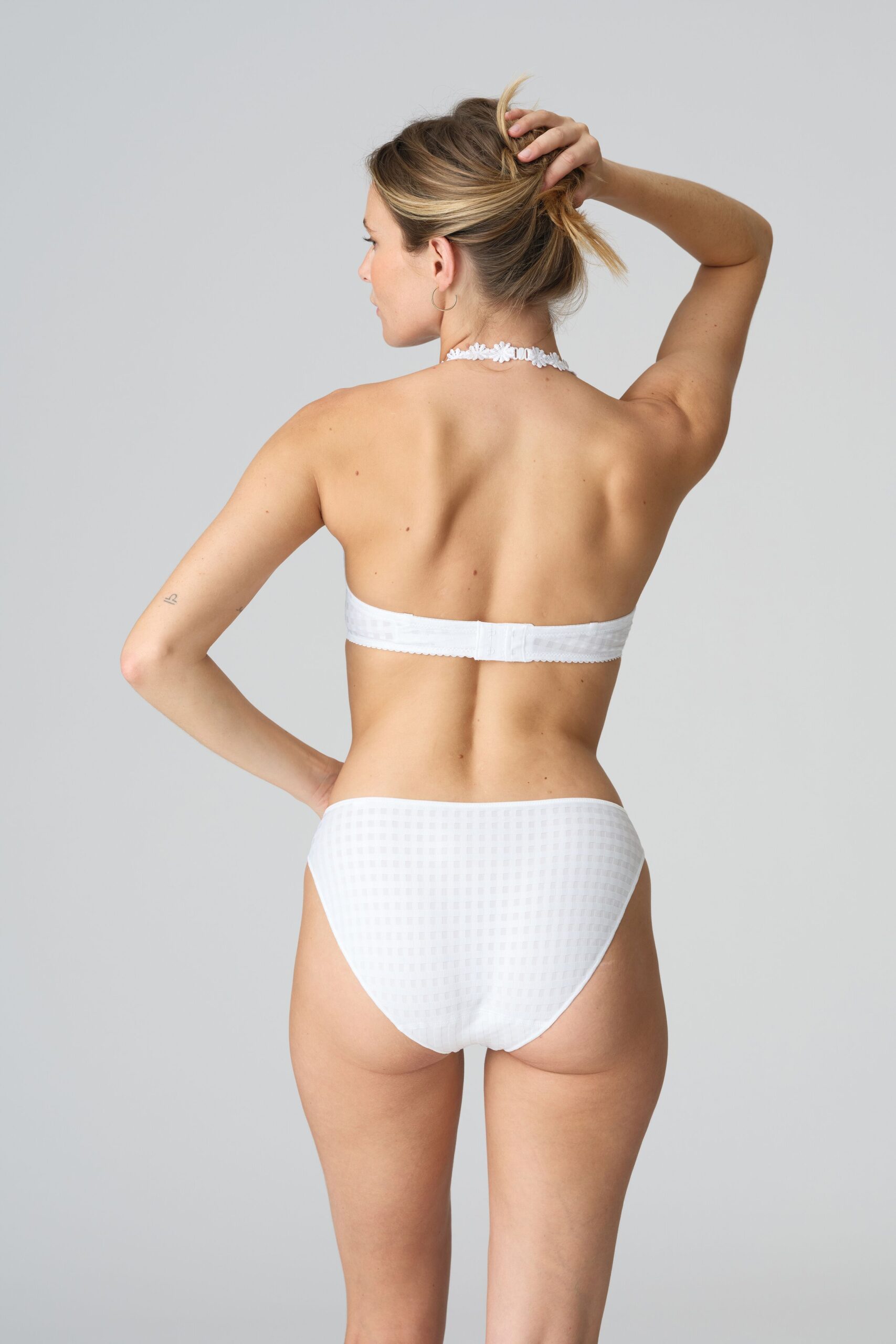 Back view of a woman wearing white underwear, one arm raised to touch her hair, in a neutral studio setting.