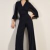 Full-body shot of a woman in a black wrap-top with flared sleeves and wide-leg pants, posing against a neutral wall.
