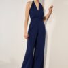 Full-length shot of a woman wearing a navy blue halter-neck jumpsuit with wide-leg pants, standing against a light wall in a studio setting.",