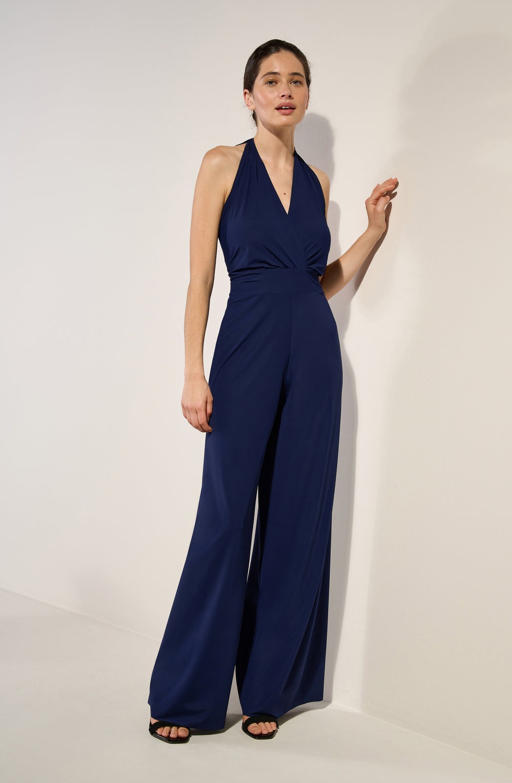 Full-length shot of a woman wearing a navy blue halter-neck jumpsuit with wide-leg pants, standing against a light wall in a studio setting.",