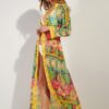 Woman in a vibrant, sheer, geometric-print cover-up over a white bikini, walking barefoot indoors beside a light wall.