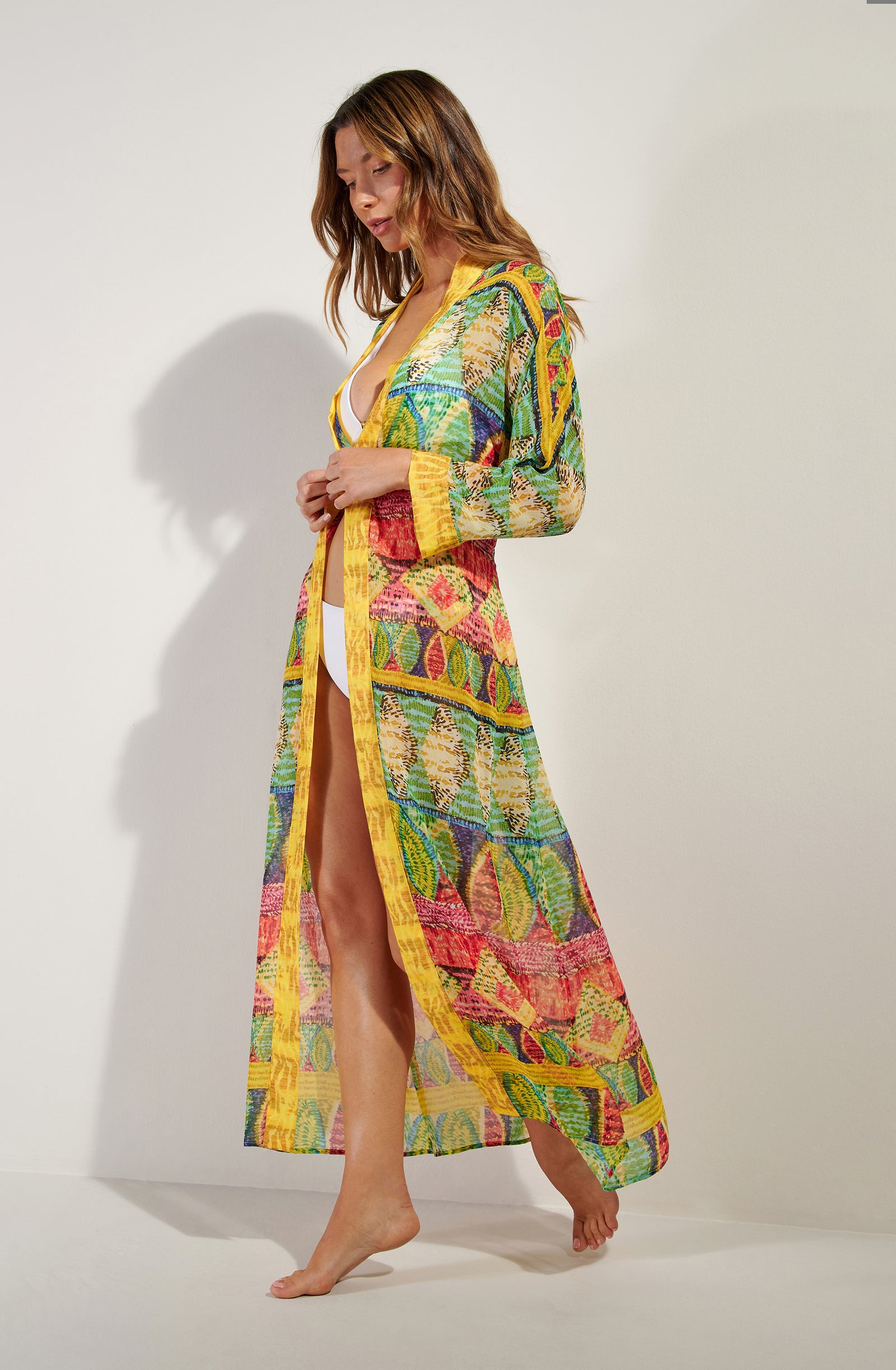 Woman in a vibrant, sheer, geometric-print cover-up over a white bikini, walking barefoot indoors beside a light wall.