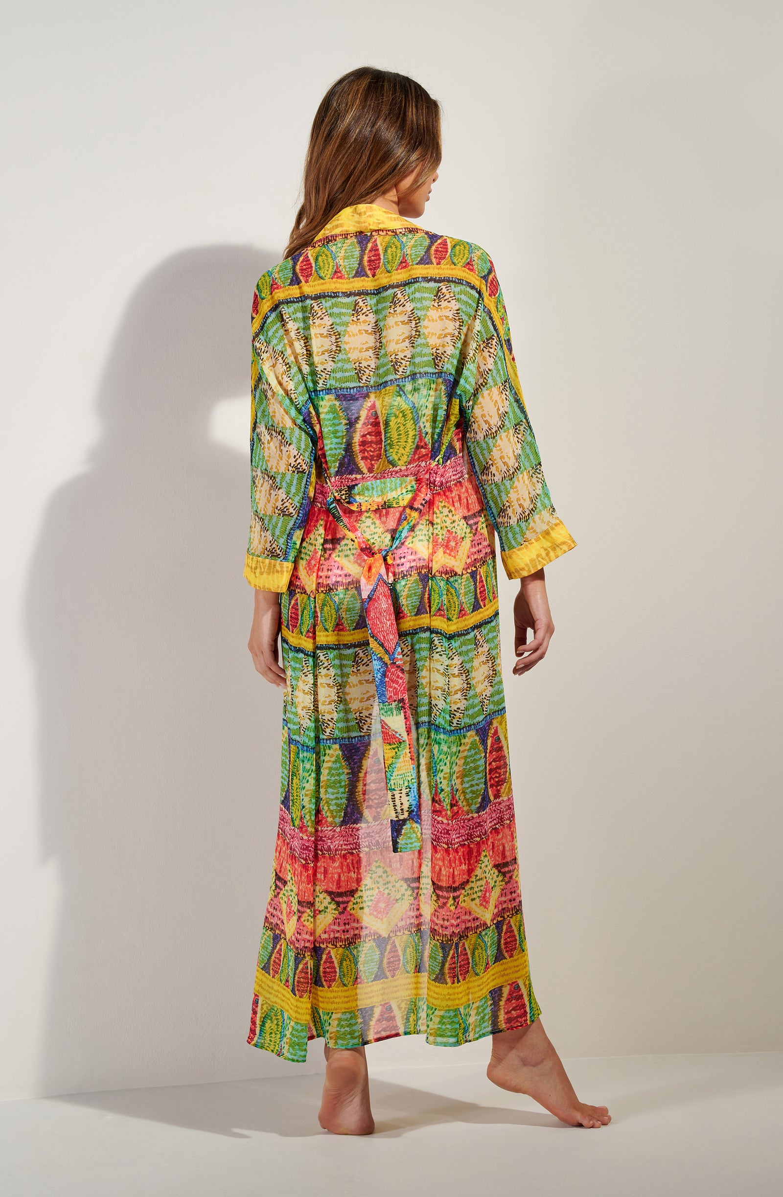 Back view of a woman wearing a bright, patterned maxi dress with yellow cuffs, standing barefoot on a white studio floor.