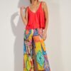 Woman wearing a bright red, layered sleeveless top and multicolored wide-leg pants with bold abstract patterns, standing against a neutral wall in a fashion pose.