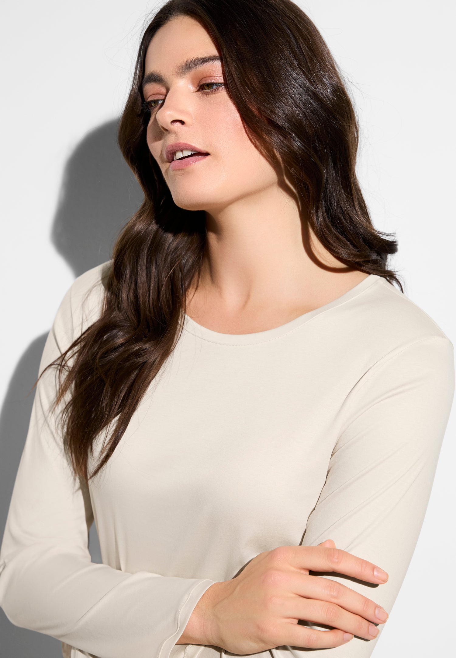 Woman with long brown hair in a beige top, arms crossed, facing slightly to the right against a light background.
