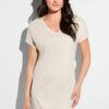 Woman wearing a light beige V-neck t-shirt dress, standing against a white wall with a shadow on the left.