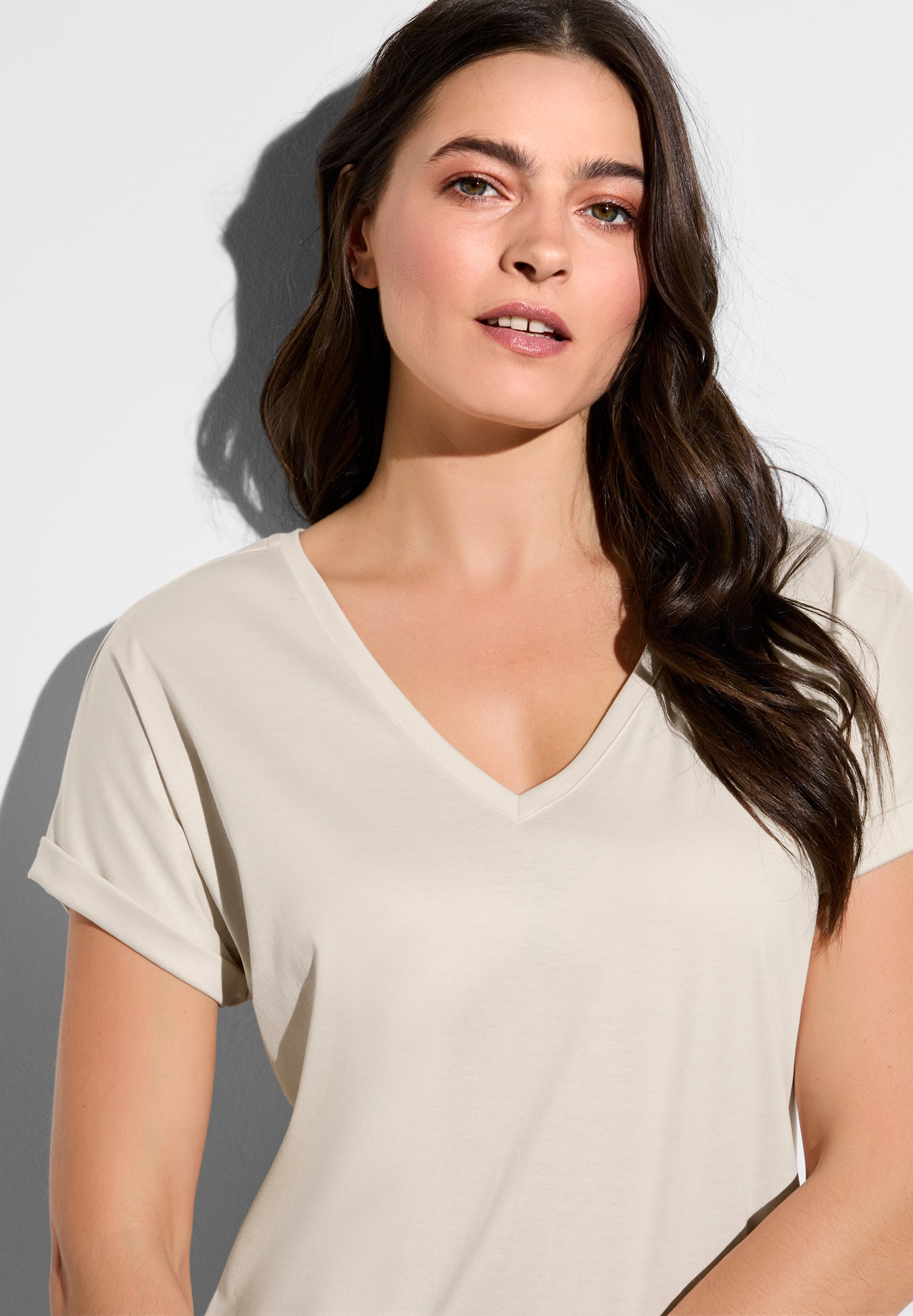 Close-up portrait of a woman with long brown hair wearing a beige V-neck shirt against a gray background.