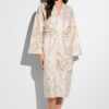 Woman standing in a cream paisley robe, facing the camera against a white background, bare feet visible.